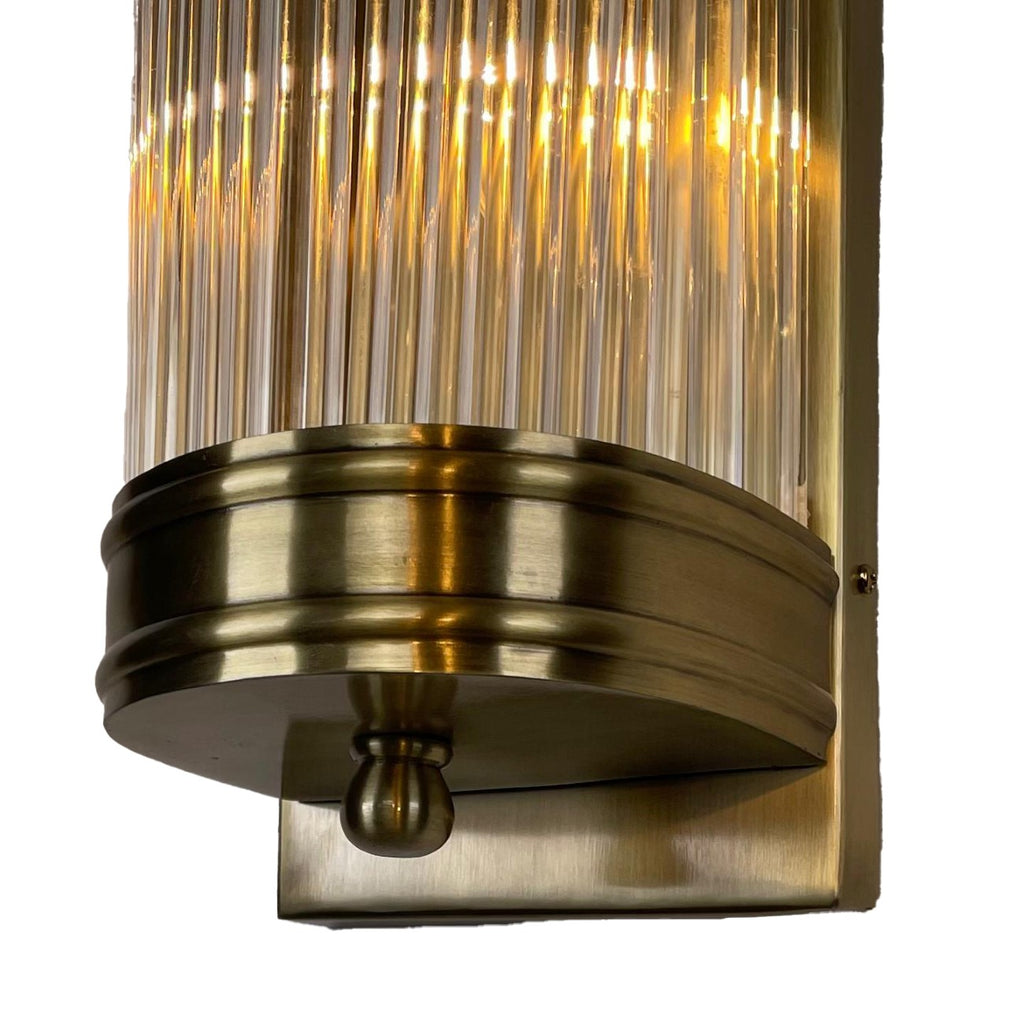 Close-up of the illuminated glass rods above the wall light's lower antique-brass-finished band, rounded finial and rectangular backplate.