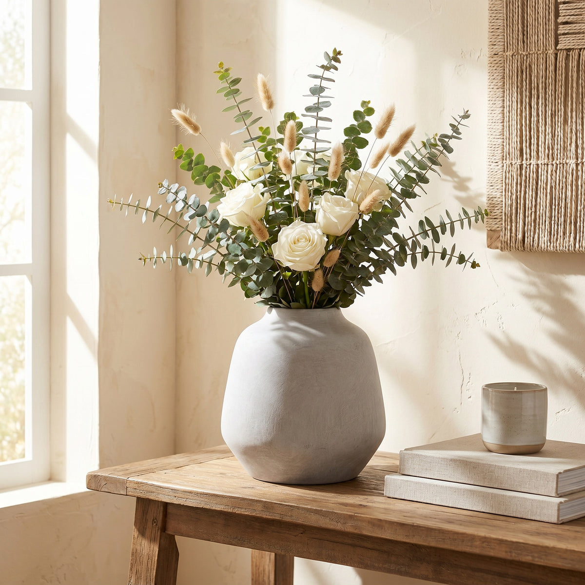 Rounded pale vase styled with white roses, leafy stems and beige grasses on a wooden console beside stacked books and a candle.