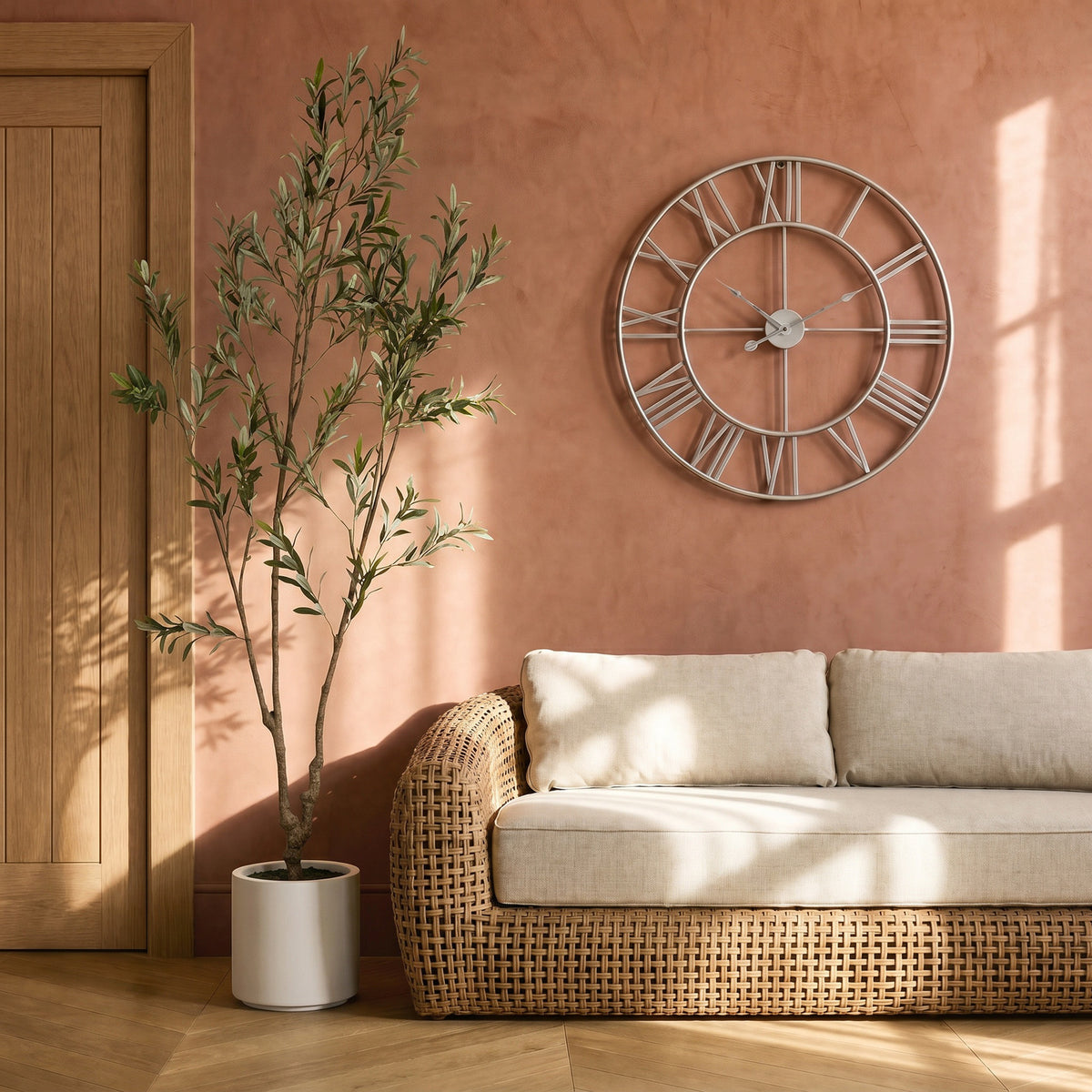 Silver-toned Roman Orbit wall clock on a terracotta-coloured wall above a woven sofa with cream cushions, beside a potted tree and wooden door.