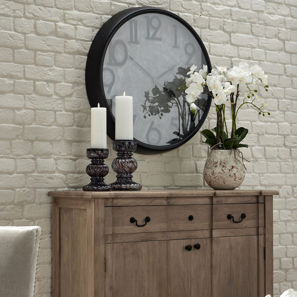 White orchid arrangement in a mottled off-white pot on a wooden sideboard, beside two tall candles beneath a large black-framed wall clock.