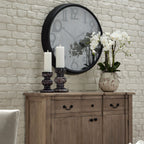 White orchid arrangement in a mottled off-white pot on a wooden sideboard, beside two tall candles beneath a large black-framed wall clock.