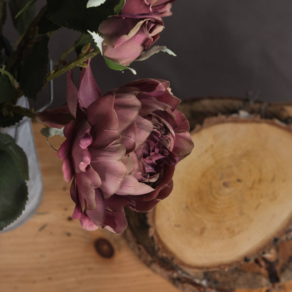 Close-up of layered dusty-pink petals and a closed bud beside dark green foliage, with part of a glass vase and a wood slice in the background.