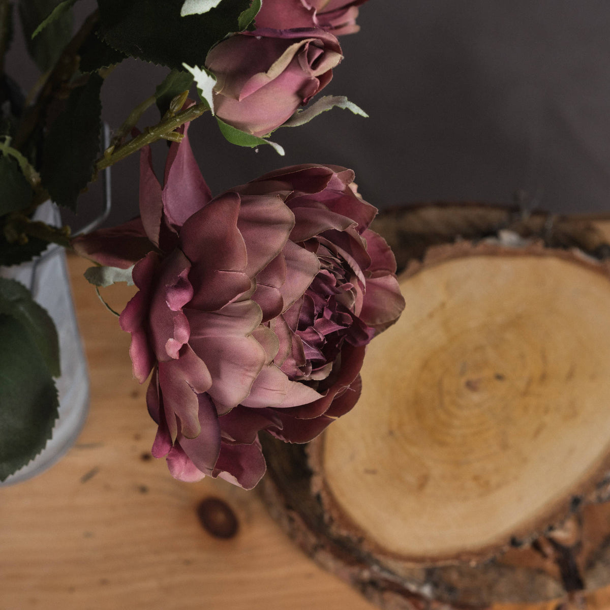 Close-up of layered dusty-pink petals and a closed bud beside dark green foliage, with part of a glass vase and a wood slice in the background.