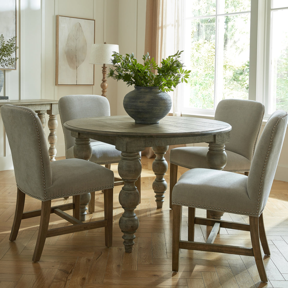 Round dining table styled with pale upholstered chairs and a dark vase of green foliage beside tall windows.