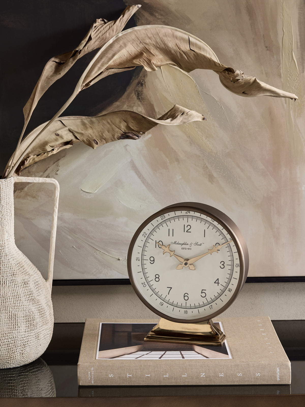 Pedestal mantel clock displayed on a clothbound book beside an ivory textured vase with dried leaves, in front of neutral abstract artwork.