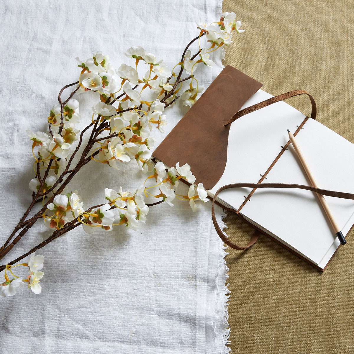 White blossom branches laid across pale cloth beside an open notebook with a brown cover, tie and pencil.