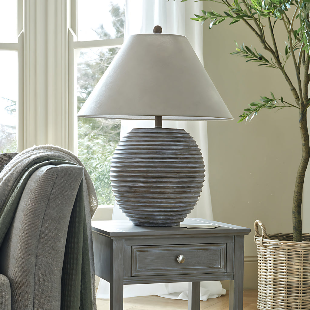 Ribbed brown table lamp on a grey side table beside an armchair with layered throws and a leafy tree in a basket.