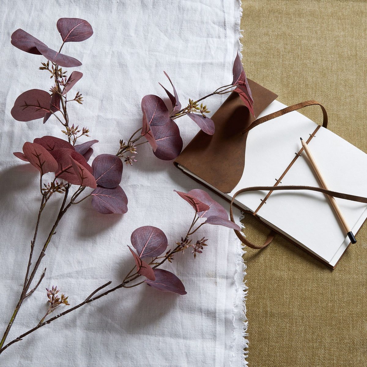 Burgundy artificial foliage branches spread across white cloth beside an open notebook and pencil on a tan textured surface.