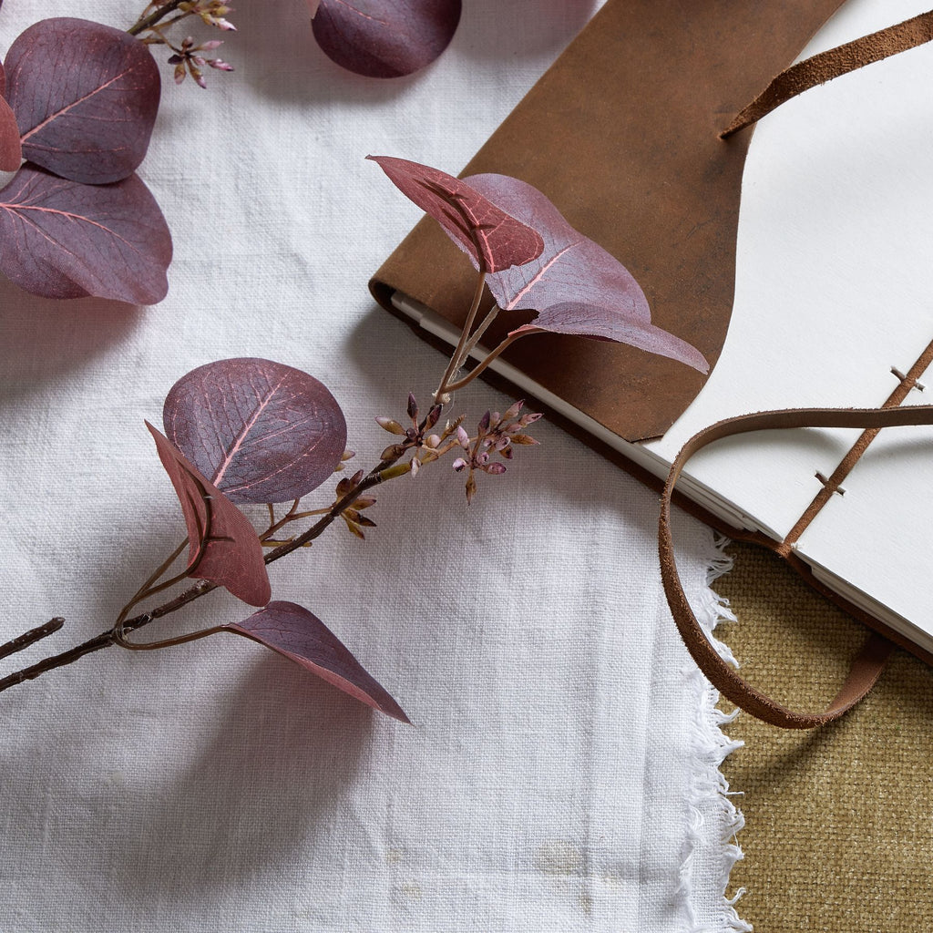 Close-up of burgundy leaves with lighter vein markings and small bud-like clusters resting on white cloth beside a brown-covered notebook.
