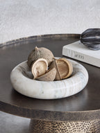 Beige marble bowl holding dried seed pods on a dark round table beside a white book and dark ornament.
