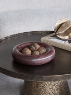 Red marble bowl filled with textured dried seed pods on a dark round table beside a book and a gold-coloured sculpture.