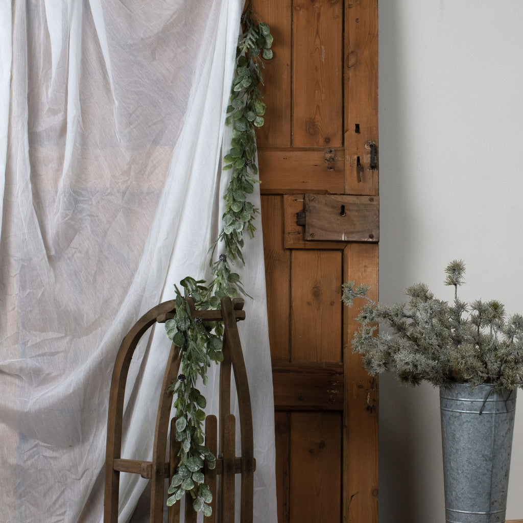 Green foliage garland hanging beside white fabric and trailing over a curved wooden frame, with a wooden door and a metal bucket of foliage nearby.