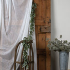 Green foliage garland hanging beside white fabric and trailing over a curved wooden frame, with a wooden door and a metal bucket of foliage nearby.