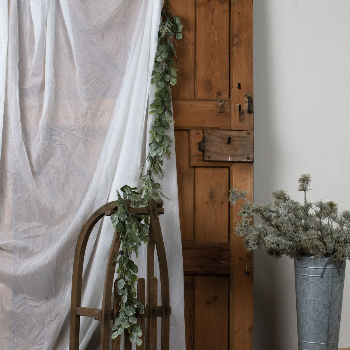 Green foliage garland hanging beside white fabric and trailing over a curved wooden frame, with a wooden door and a metal bucket of foliage nearby.