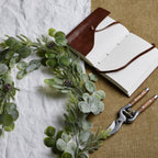 Frosted-looking foliage garland laid across white and tan fabric beside an open notebook with a brown cover and pruning shears.