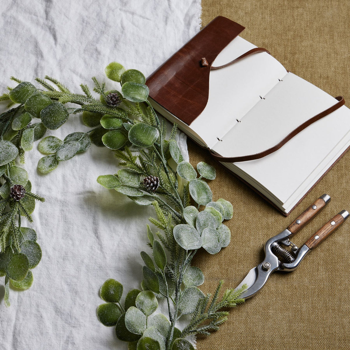 Frosted-looking foliage garland laid across white and tan fabric beside an open notebook with a brown cover and pruning shears.