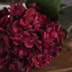 Close-up of overlapping magenta and wine-toned hydrangea petals, showing curled edges and small flower centres with green foliage behind.