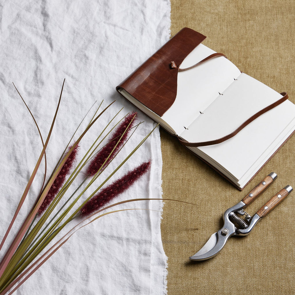 Ruby Plume spray laid diagonally across white fabric beside an open brown-covered notebook and secateurs on a tan woven surface.