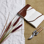 Ruby Plume spray laid diagonally across white fabric beside an open brown-covered notebook and secateurs on a tan woven surface.