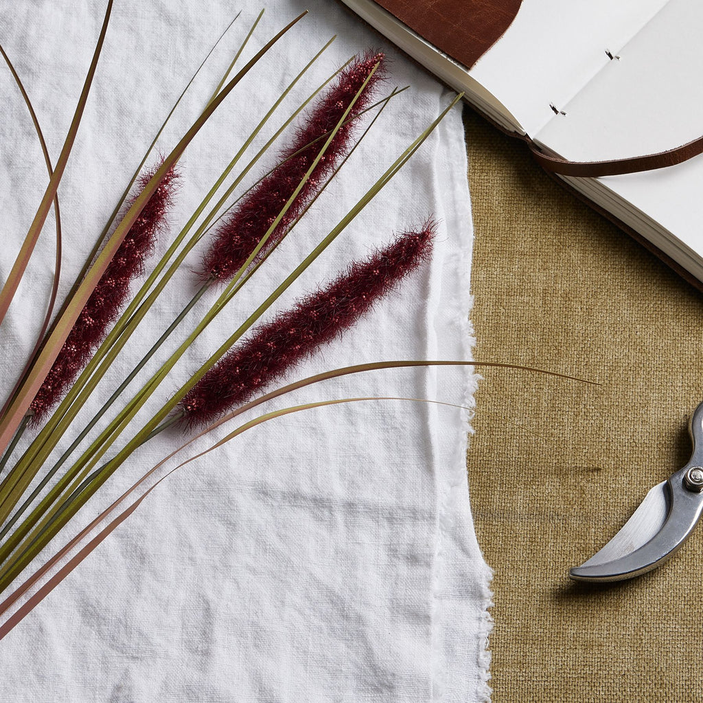 Close-up of three fuzzy ruby plumes crossed by narrow green and reddish blades on white fabric, beside the edges of an open notebook and secateurs.