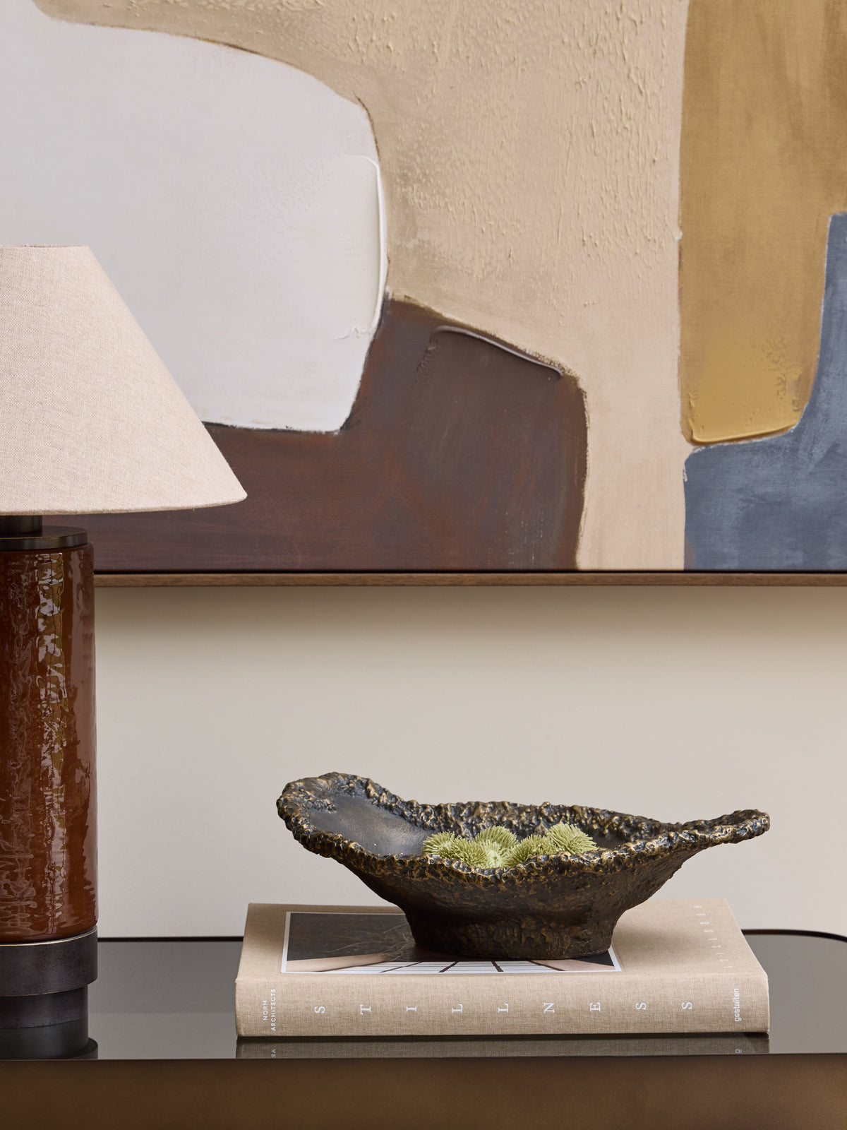 Dark bronze bowl holding small green decorative stems on a pale book, beside a brown table lamp beneath abstract artwork.