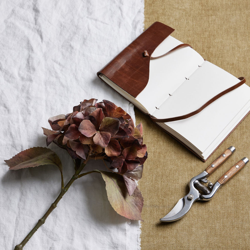 Russet artificial flower stem laid diagonally across white cloth, with an open brown-covered notebook and wooden-handled secateurs on a tan surface.