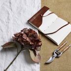 Russet artificial flower stem laid diagonally across white cloth, with an open brown-covered notebook and wooden-handled secateurs on a tan surface.