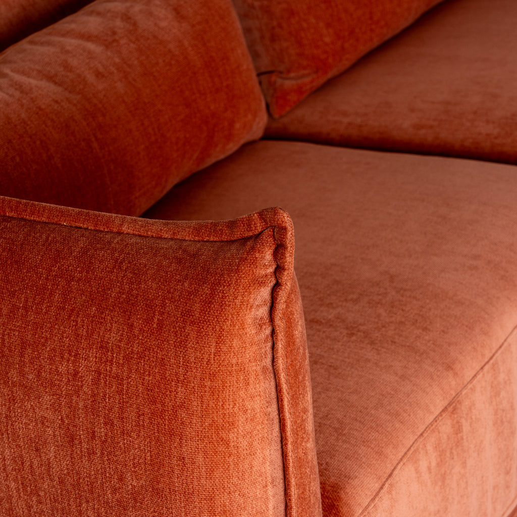 View over a rounded sofa arm highlighting the raised edge seam, with rust-orange seat and back cushions behind.