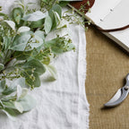 Close-up of softly textured sage-green leaves and tiny white flowers or buds on fine branches, laid on white cloth beside part of an open notebook and secateurs.