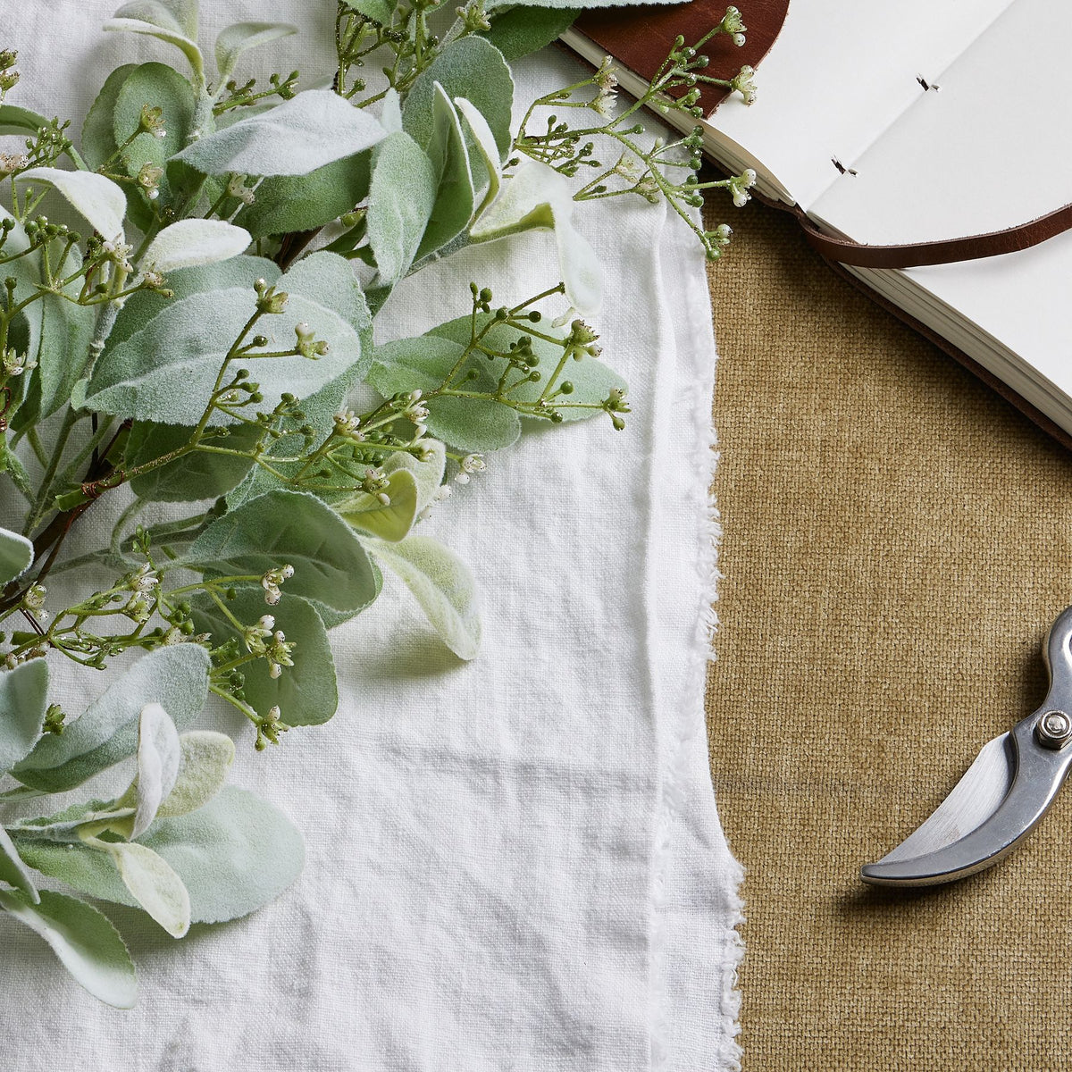 Close-up of softly textured sage-green leaves and tiny white flowers or buds on fine branches, laid on white cloth beside part of an open notebook and secateurs.