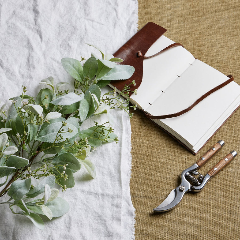 Sageleaf artificial foliage laid diagonally across white cloth beside an open brown-covered notebook and secateurs on a tan woven surface.
