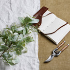 Sageleaf artificial foliage laid diagonally across white cloth beside an open brown-covered notebook and secateurs on a tan woven surface.
