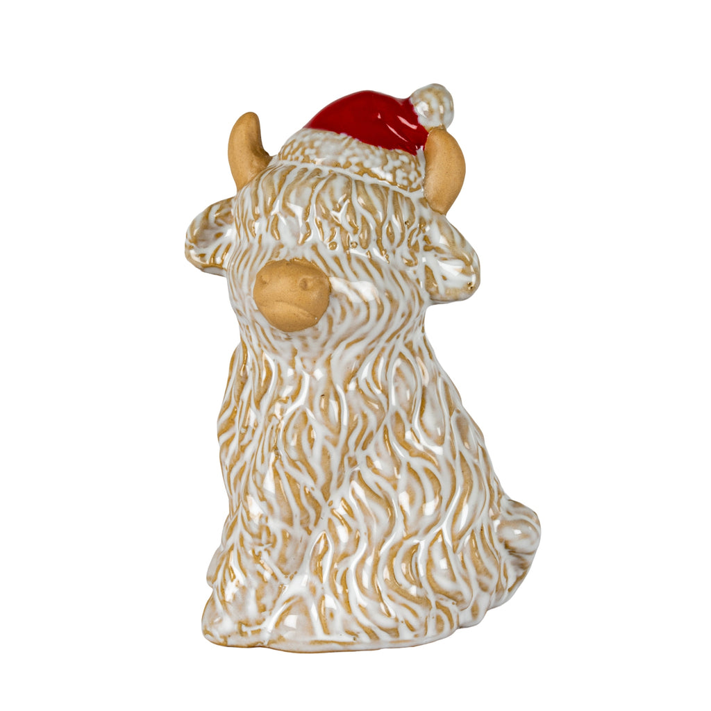 Full view of a seated cream-and-tan shaggy cow decoration wearing a red Santa-style hat, with tan horns, folded front legs and a small tail visible.