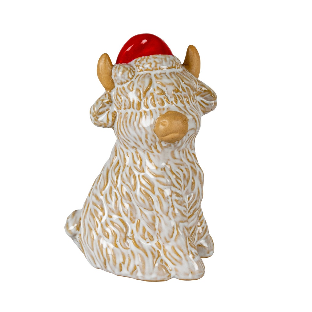 Seated cream-and-tan shaggy cow figure wearing a red Santa hat, with tan horns, a rounded muzzle, folded front legs and a small tail visible at the side.