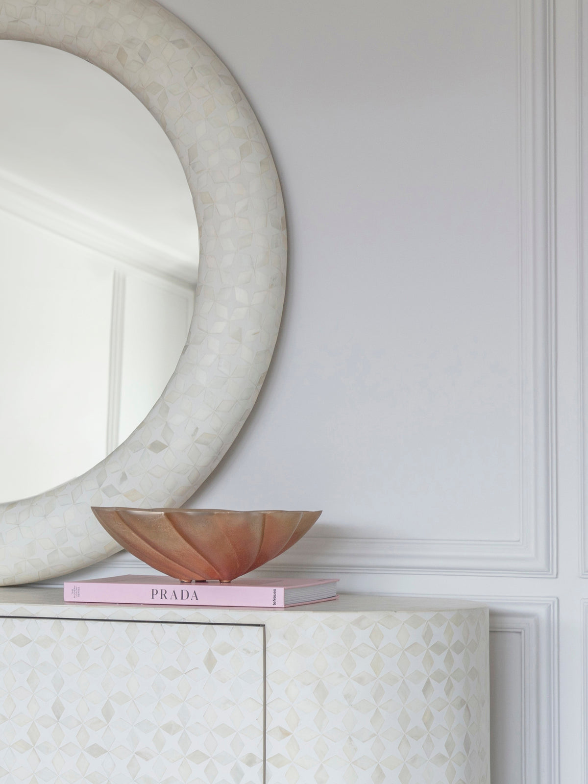 Gold decorative bowl on a pink book atop a pale patterned cabinet beneath a round mirror.