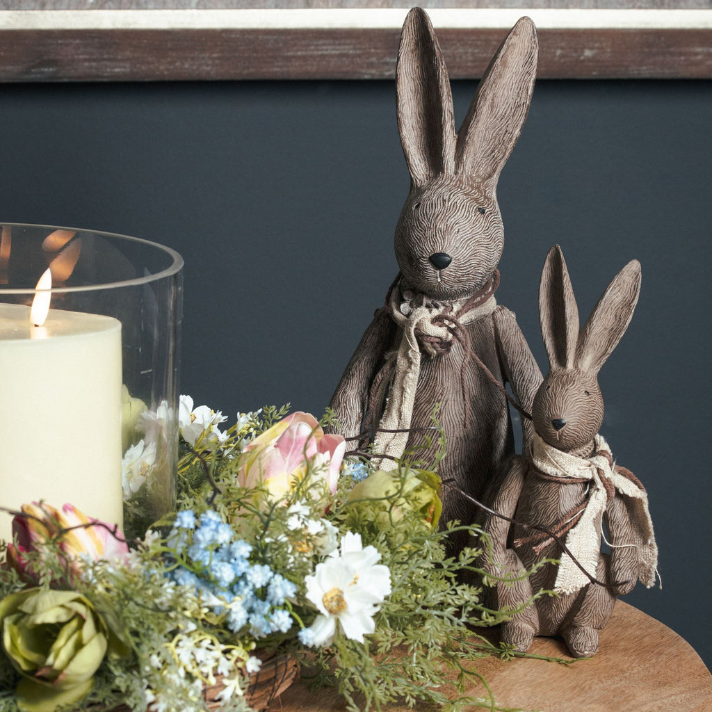 Two scarf-wearing rabbit ornaments on a wooden table beside flowers and a candle in glass, with the larger rabbit standing behind the smaller one.