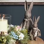 Two scarf-wearing rabbit ornaments on a wooden table beside flowers and a candle in glass, with the larger rabbit standing behind the smaller one.