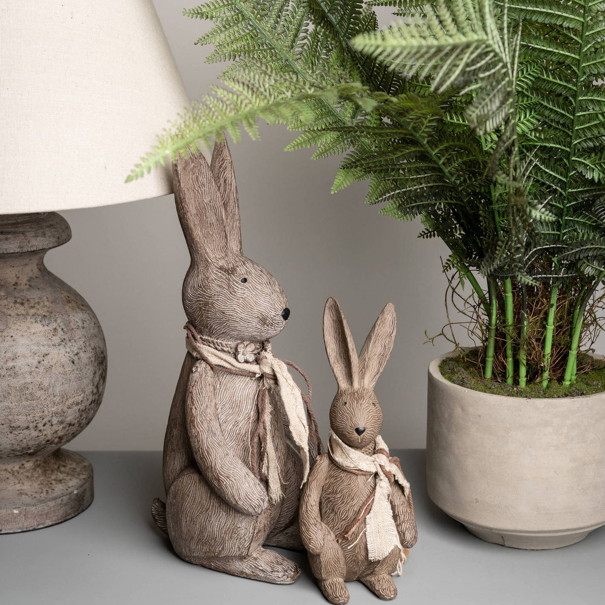 Small rabbit ornament beside a larger matching rabbit on a grey surface, between a table lamp and a potted fern.