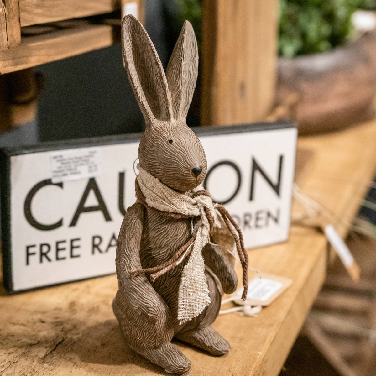 Scarf-wearing rabbit ornament on a wooden shelf in front of a black and white sign, with its long ears and dark nose clearly visible.