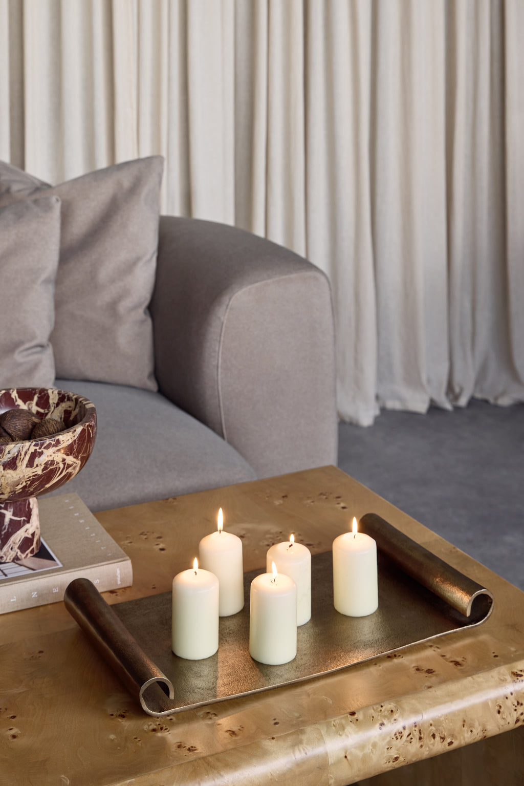 Scroll-end tray holding five lit cream pillar candles on a wooden coffee table beside books and a patterned bowl, with a grey sofa behind.