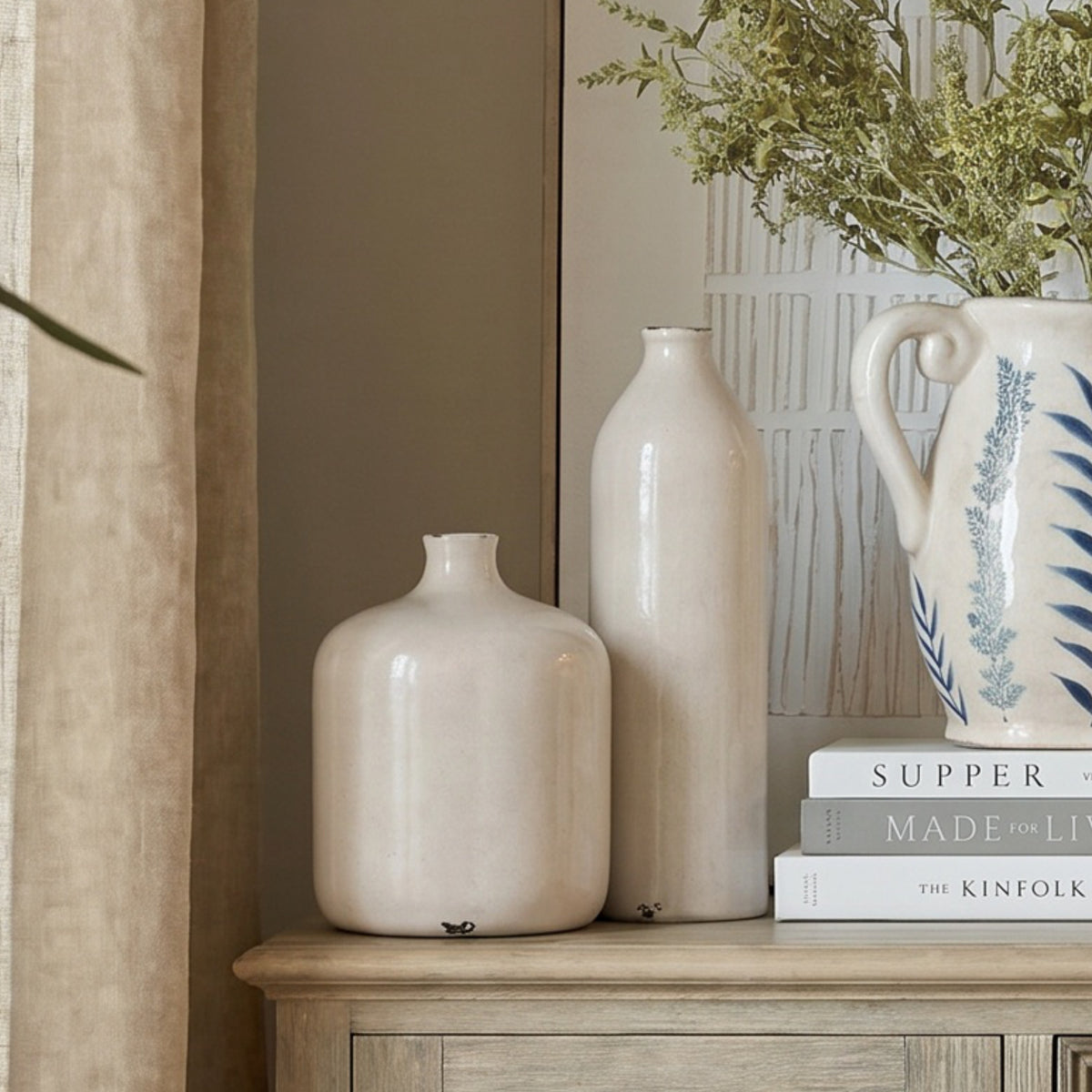 Tall off-white vase on a pale wooden cabinet beside a shorter rounded vase and a blue-patterned jug of leafy stems on stacked books.