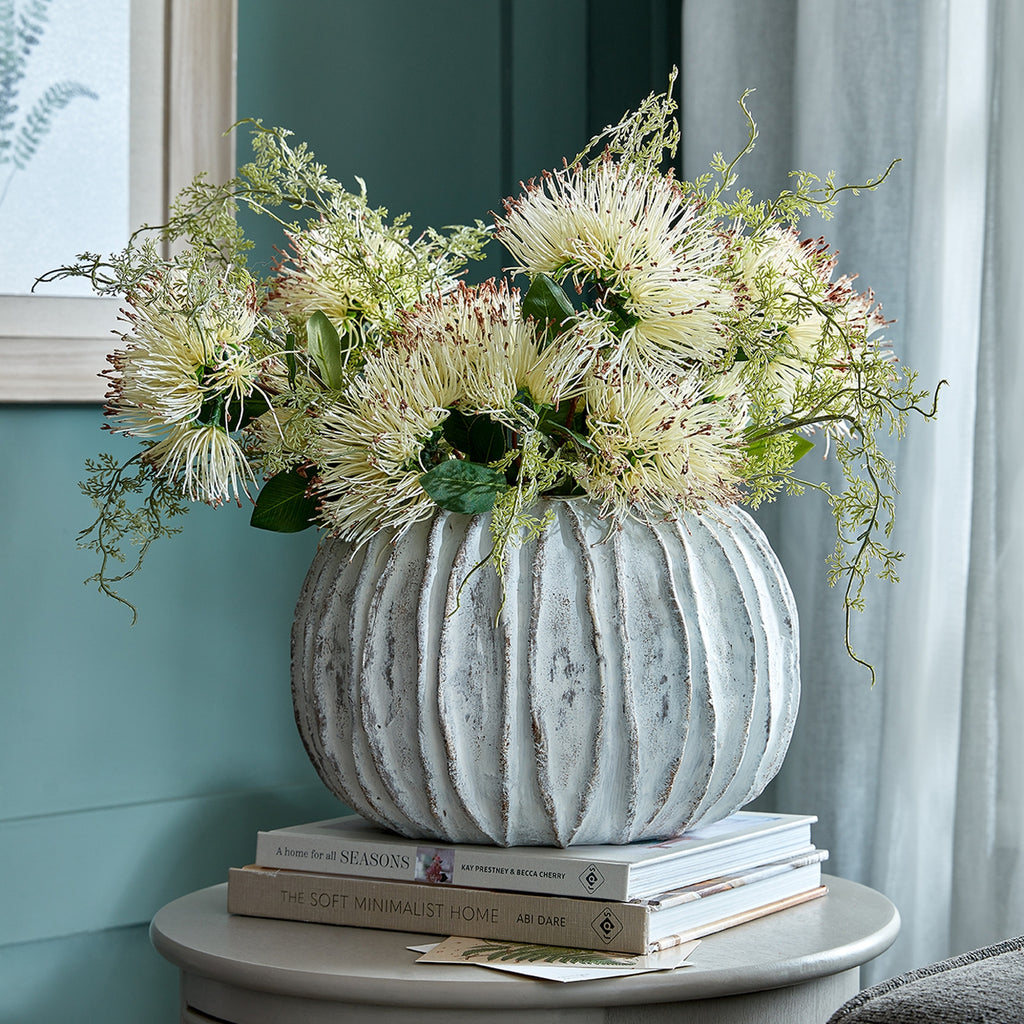 Pale ribbed vase filled with cream flowers and trailing greenery on stacked books, beside a teal wall and light curtain.