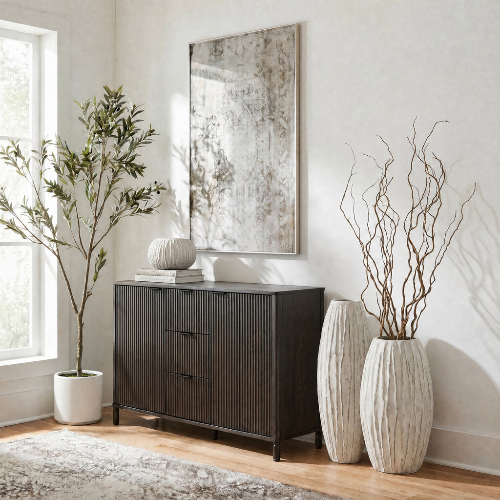 Pale ribbed vases beside a dark fluted cabinet, with tall twisting branches in the front vase and framed artwork above.