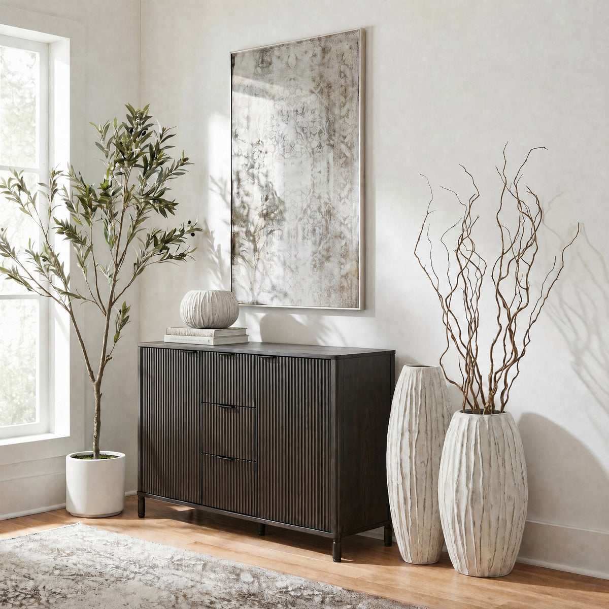 Pale ribbed vases beside a dark fluted cabinet, with tall twisting branches in the front vase and framed artwork above.