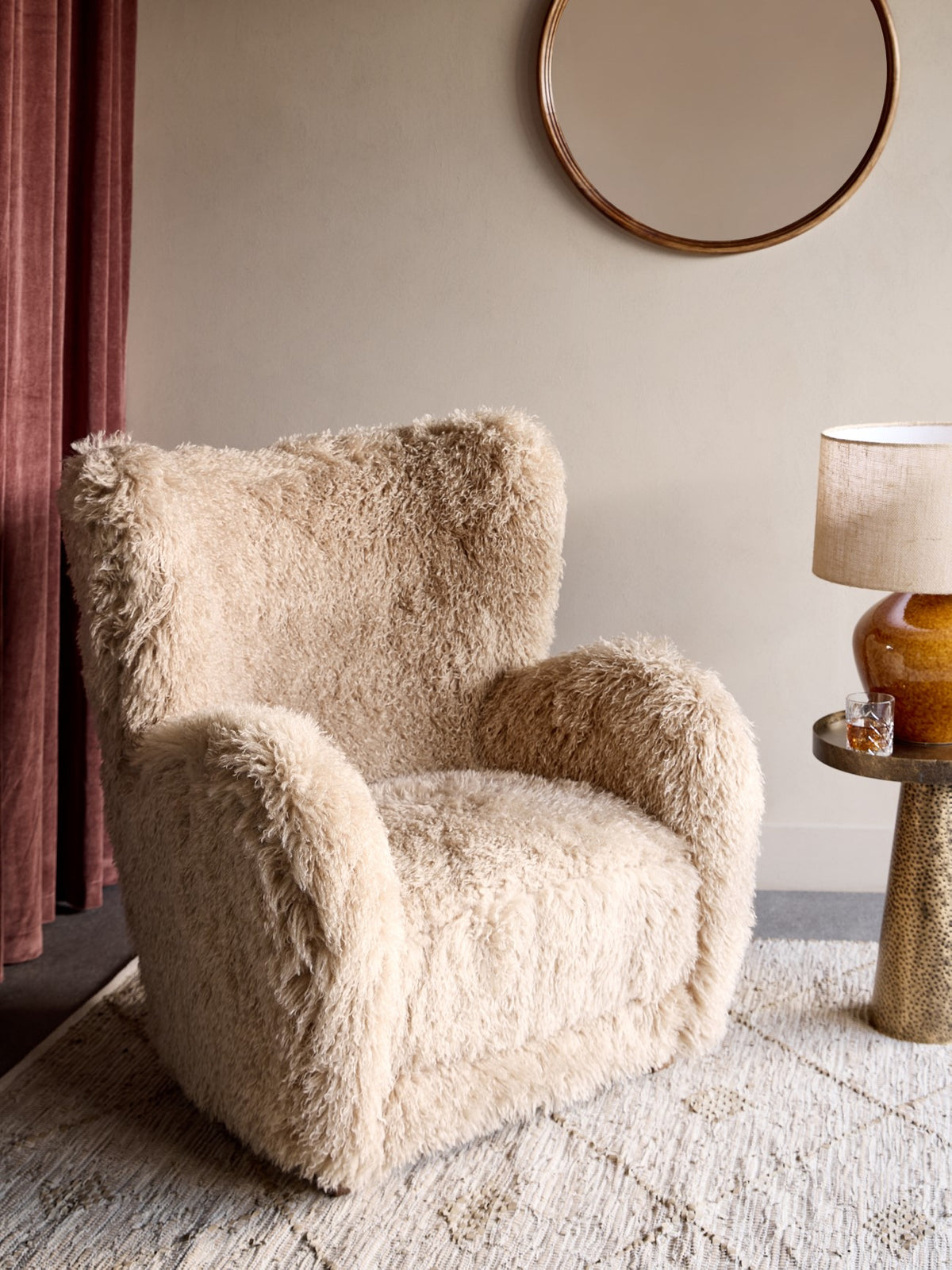 Shaggy cream armchair on a pale patterned rug beside rust-coloured curtains and a side table with an amber lamp.