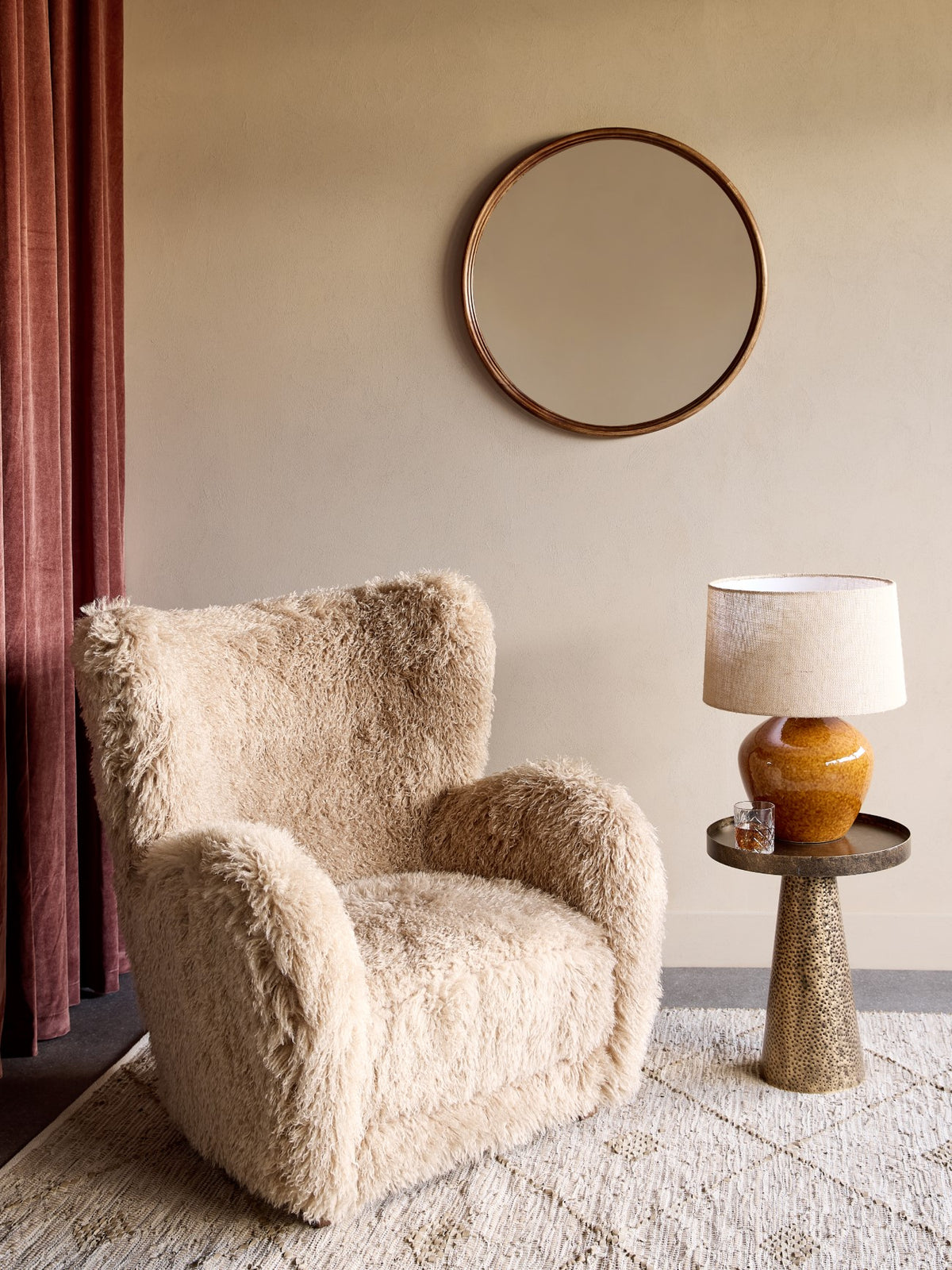 Cream shaggy armchair in a room setting with a round wall mirror, rust curtains and a metal side table holding an amber lamp.