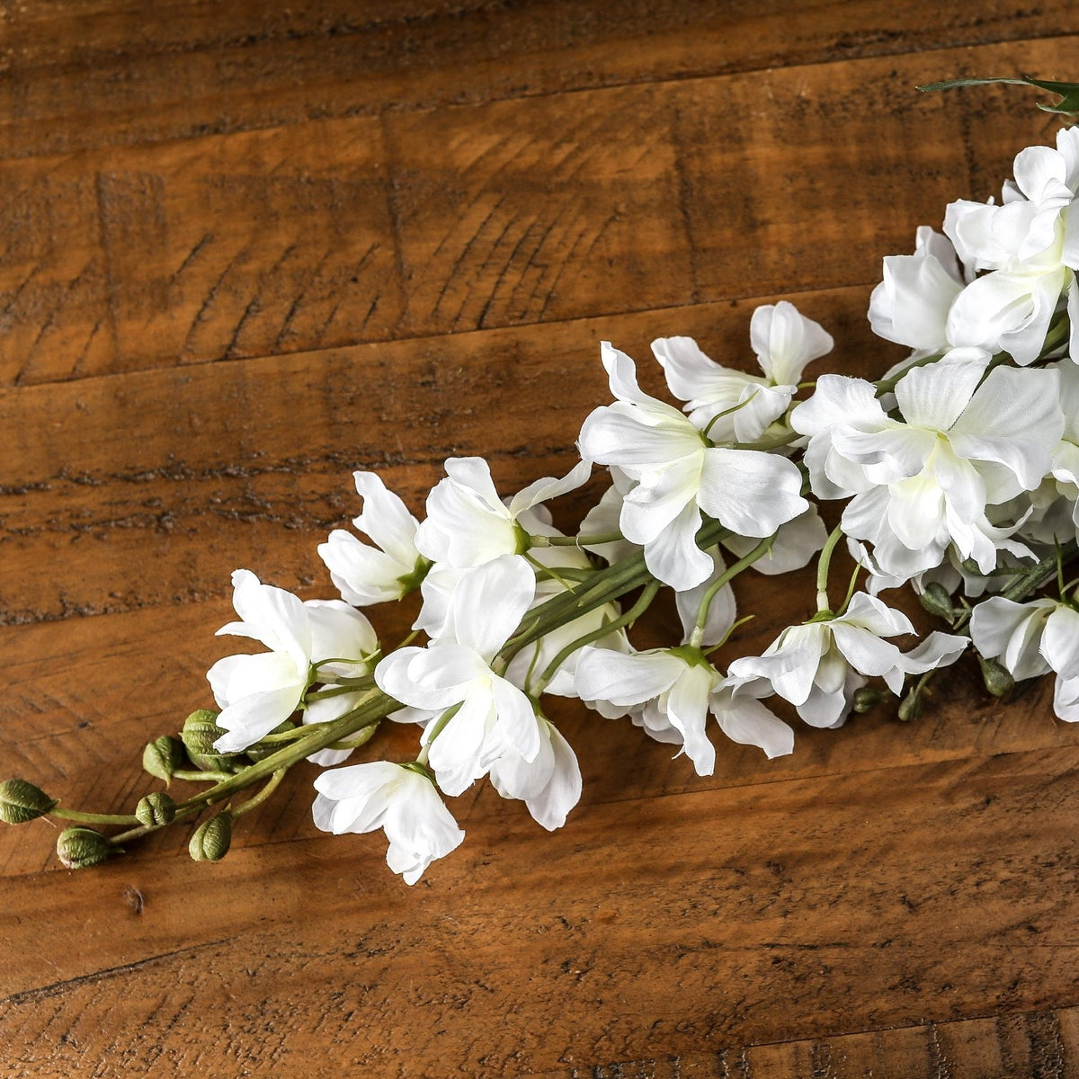 Close-up of white artificial florets with pale centres and green buds, lying diagonally across a brown wooden surface.