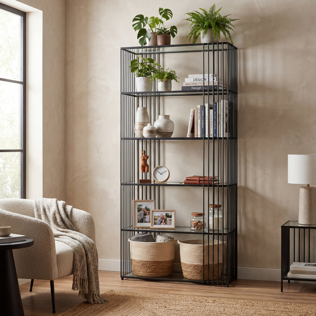 Black shelving unit beside an armchair in a pale living room, styled with plants, books, vases, framed photographs and woven baskets.