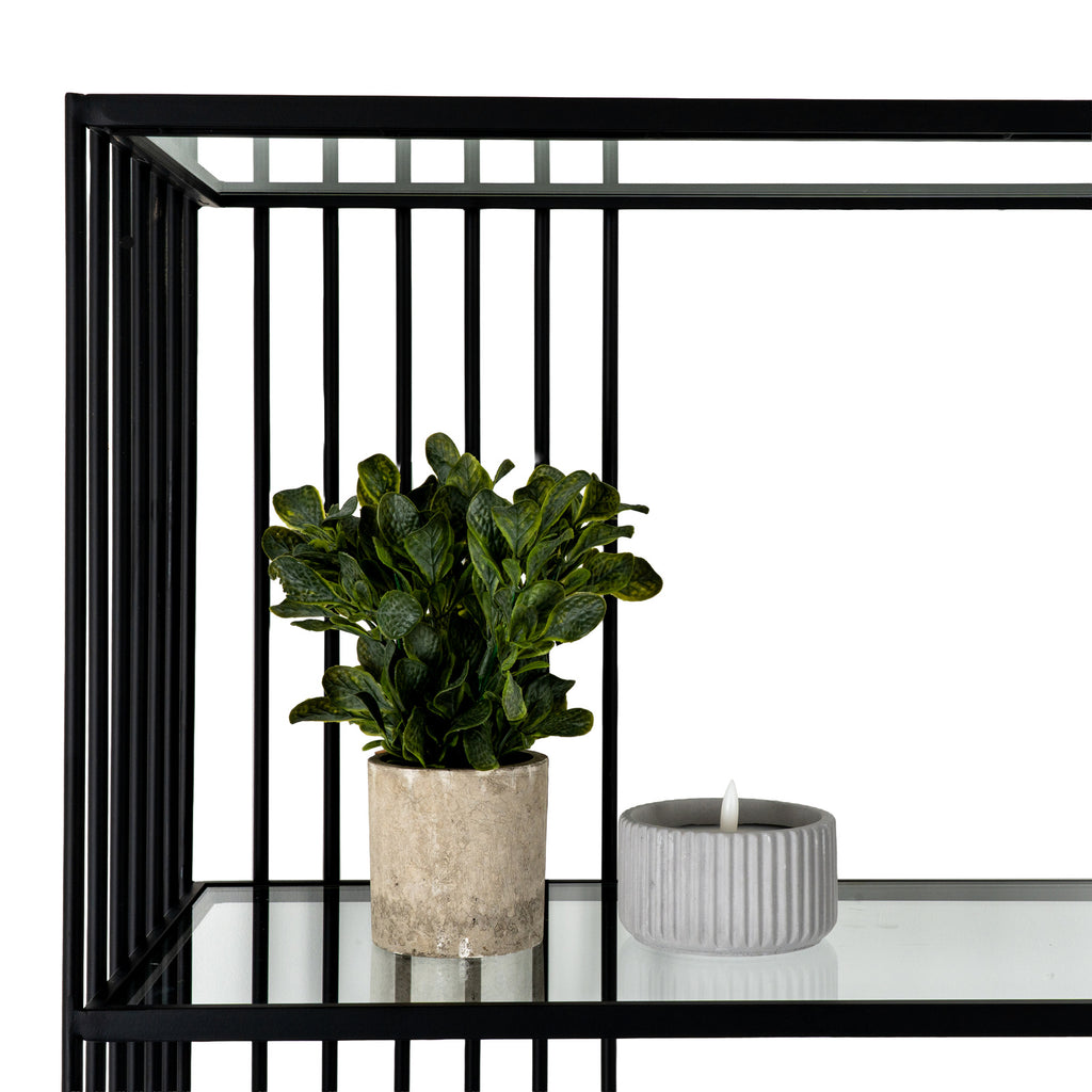 Close-up of a clear shelf beneath black vertical rods, styled with a leafy plant in a beige pot and a grey ribbed candle ornament.