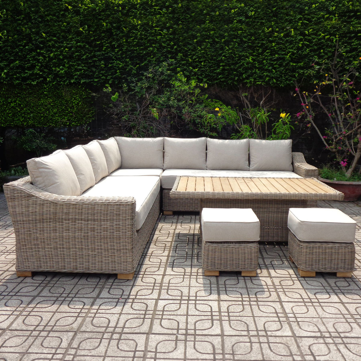 Pale woven-look corner seating with cream cushions, a rectangular slatted-top table and two square cushioned stools on patterned paving against dense greenery.